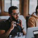Konzentrierter Mann passt Kopfhörer an, während er am Laptop arbeitet, Kollege im Hintergrund. erecruiter, Bewerbermanagement